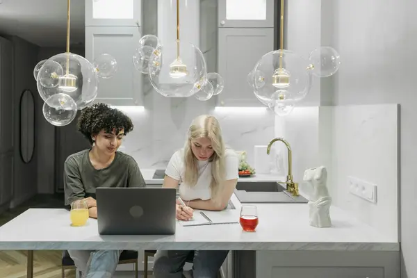A laptop and notebook open on a sunlit kitchen table with people gathered around it talking